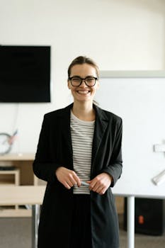 Caucasian woman in black blazer with eyeglasses, smiling in classroom.