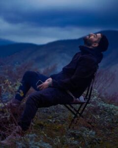 A man enjoys a serene moment outdoors, seated on a chair amidst nature under the twilight sky.