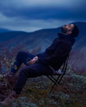A man enjoys a serene moment outdoors, seated on a chair amidst nature under the twilight sky.