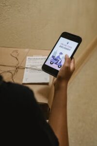 A person holding a phone displaying an online food delivery app next to a pizza box indoors.